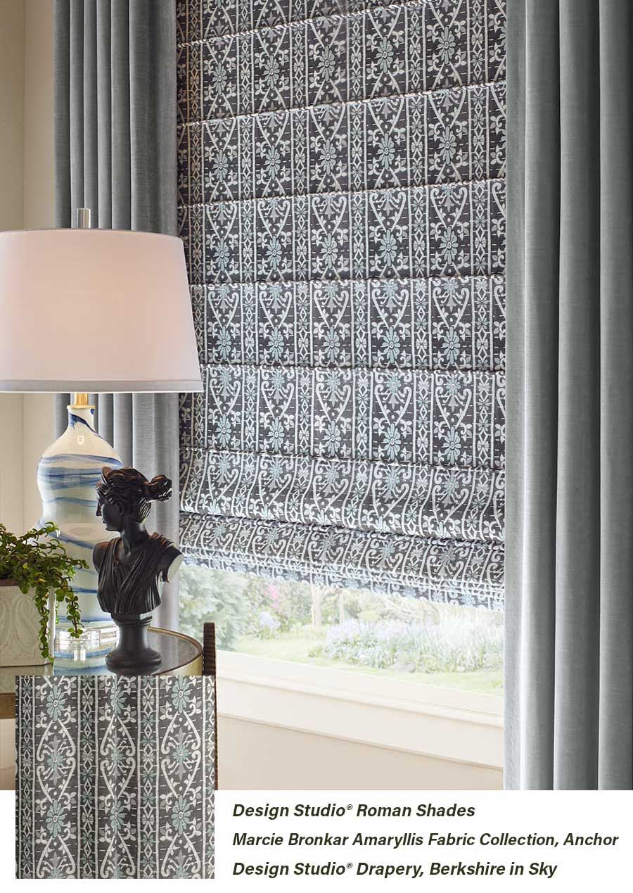 An area of a living room with patterned base shades and colorful, solid draperies. It is seen with a side table lamp, neutral walls and and window.