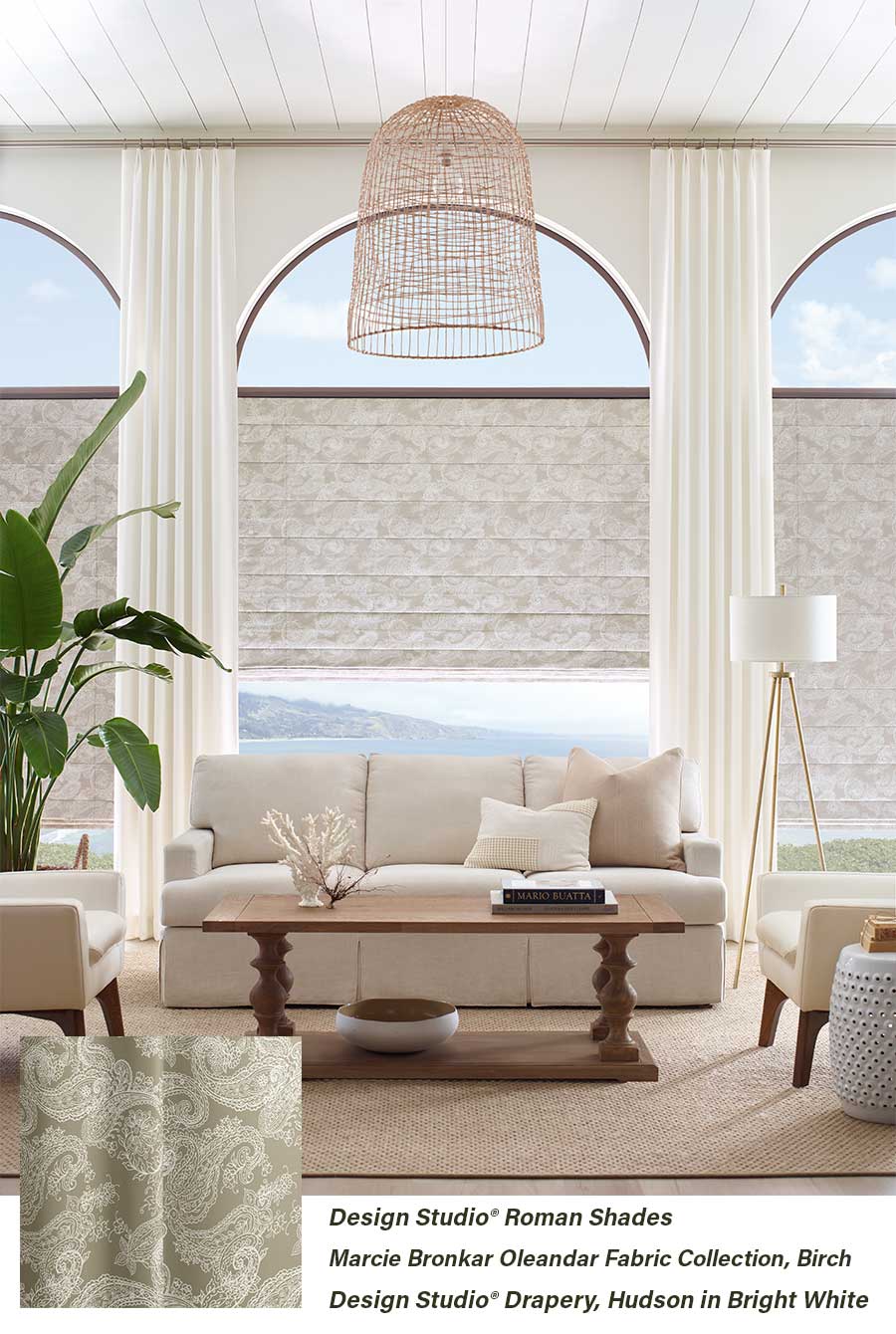 Layered ceiling height neutral colored draperies over arched windows covered in neutral roman shades in a living room setting.